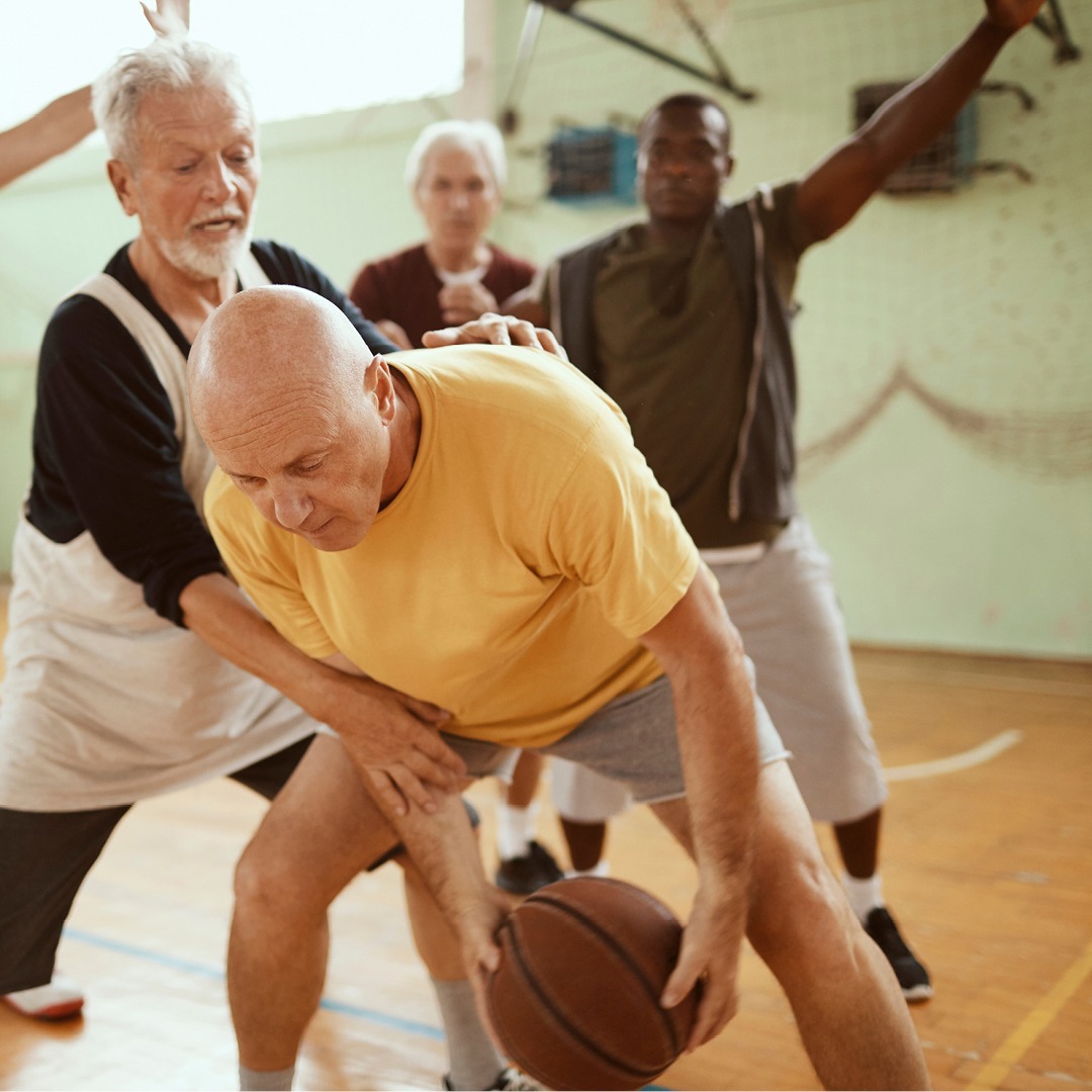 Active older adult men playing basketball in gymnasium.